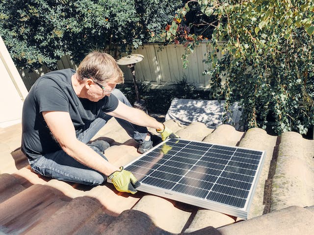 A Man in Black Shirt Sitting on the Roof while Holding a Solar Technology
