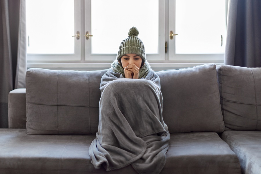 A woman is sitting on a couch while she’s wearing a hat and has a blanket thrown over her.