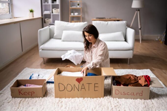 A woman sits in her living room, organizing clothes into three boxes labeled "Keep," "Donate," and "Discard."