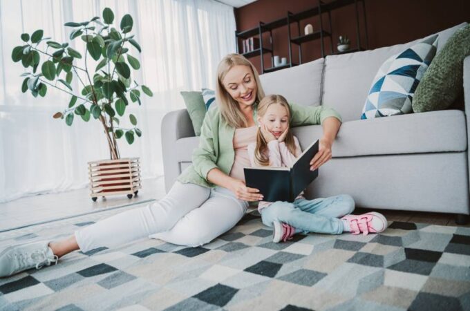 A mom sitting on the floor with her daughter, reading a book. They are on a checkered rug and leaning against a sofa.