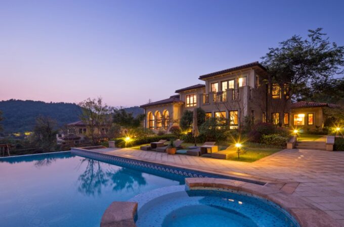 A luxurious backyard with a pool and sitting area illuminated by pathway lights. The lights are on in the house.