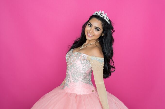 A young woman wearing a sparkly tiara and a full pink dress while standing against a hot pink background.
