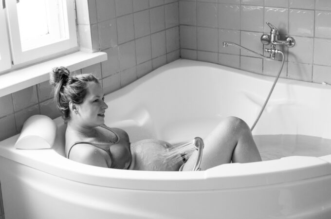 A black and white photo shows a smiling pregnant woman sitting in a tub using the handheld shower head to spray her belly.
