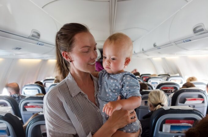 Woman holds her baby in airplane cabin aisle, smiling while passengers sit in rows of seats behind them.