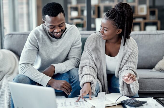 A smiling man and woman sit on a gray couch, looking at paperwork that sits on the coffee table. She's holding a pen.