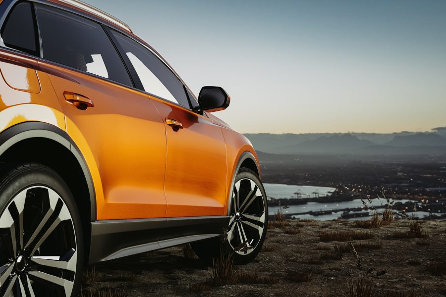 An orange SUV is parked on a grassy area overlooking a city with twinkling lights and a body of water.