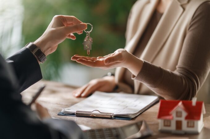 A real estate agent hands over house keys to a woman, with paperwork and a model home sitting on the desk between them.