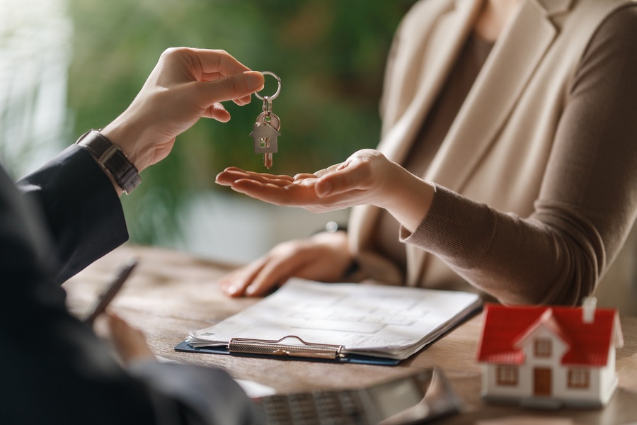 A real estate agent hands over house keys to a woman, with paperwork and a model home sitting on the desk between them.