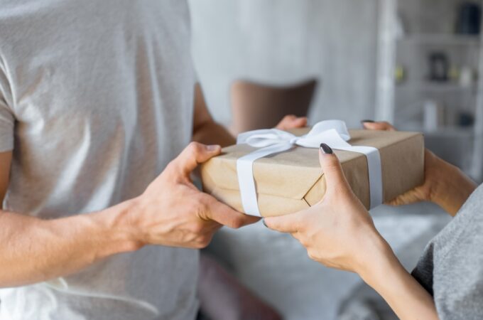 a woman handing a wrapped gift to a man in a gray shirt while standing together in a living room at home.