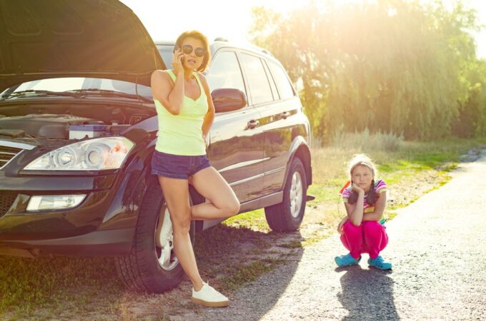 A woman stands outside her broken-down car on her cell phone. Her child crouches down next to the car on the road.