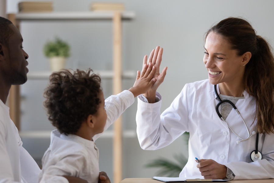 Child reaches up to high-five a smiling doctor in an exam room while an accompanying adult sits beside them.