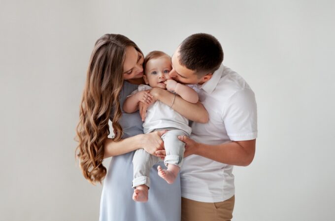 Parents kiss their baby’s cheeks while holding the child against a plain light background, with the baby looking forward.