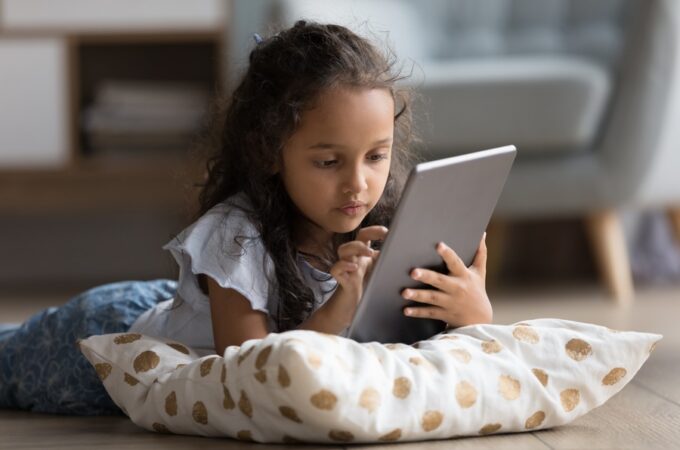 A young child lying on a wooden floor with a patterned pillow while holding and using a digital tablet indoors.