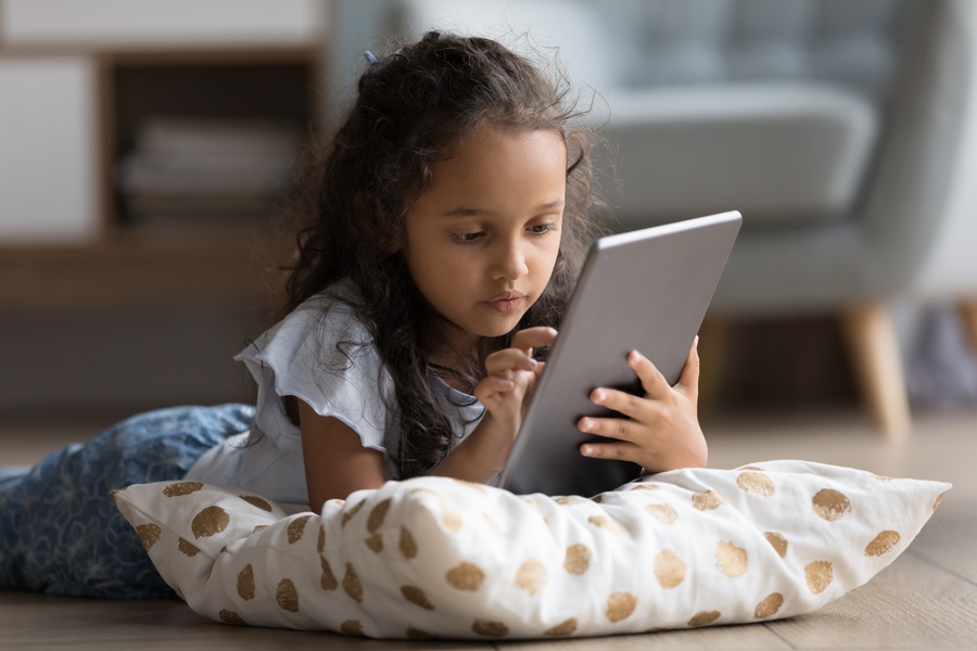 A young child lying on a wooden floor with a patterned pillow while holding and using a digital tablet indoors.