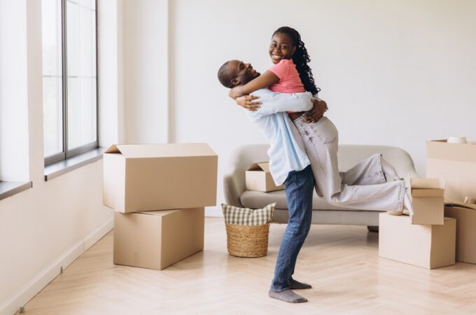 A smiling man picks up a smiling woman by the waist while standing in living room fille with moving boxes.