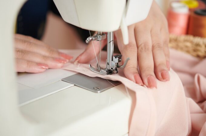 A close-up of two hands with peach nails holding a piece of pink fabric under the needle of a white sewing machine.