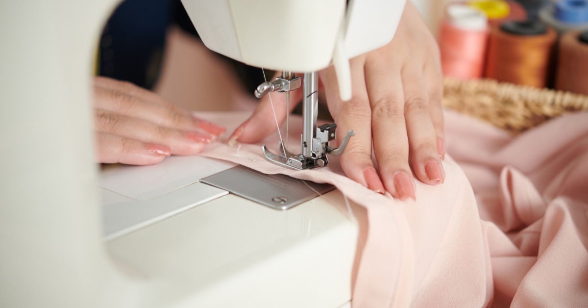 A close-up of two hands with peach nails holding a piece of pink fabric under the needle of a white sewing machine.
