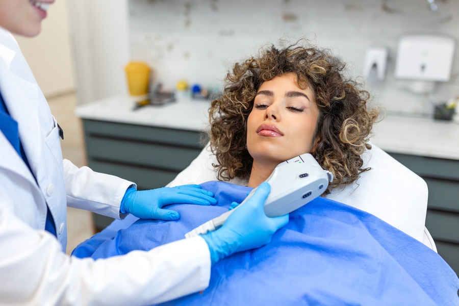 A woman is lying on a medical table. A medical professional is holding a handheld tool up to the woman's chin.