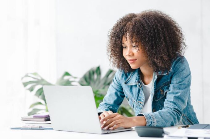 A young woman wearing a jean jacket sits at a white table typing on an open laptop. A stack of books is next to the laptop.