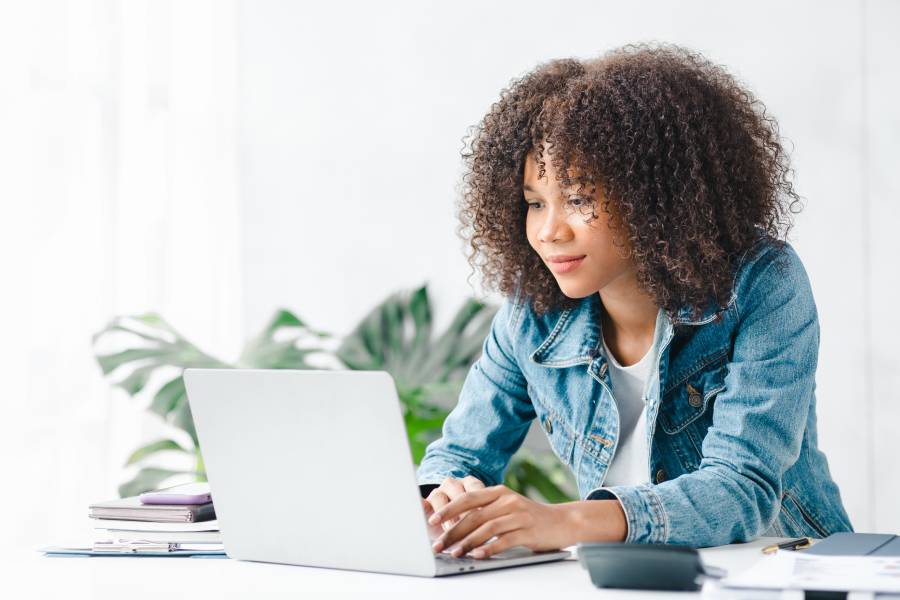 A young woman wearing a jean jacket sits at a white table typing on an open laptop. A stack of books is next to the laptop.