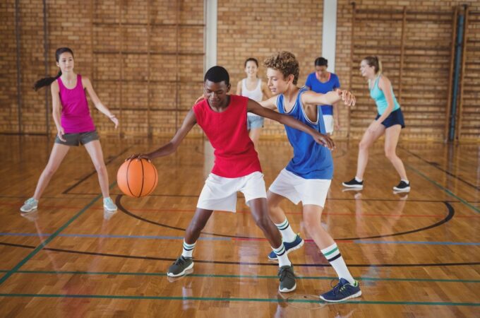 A group of 6 diverse teenagers playing basketball in a court. They are all wearing gym uniforms and shoes.