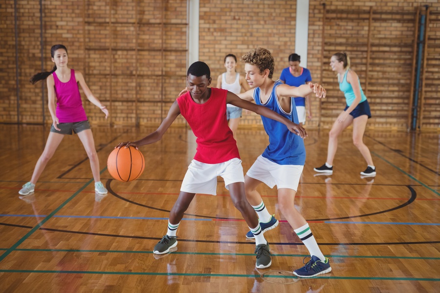 A group of 6 diverse teenagers playing basketball in a court. They are all wearing gym uniforms and shoes.