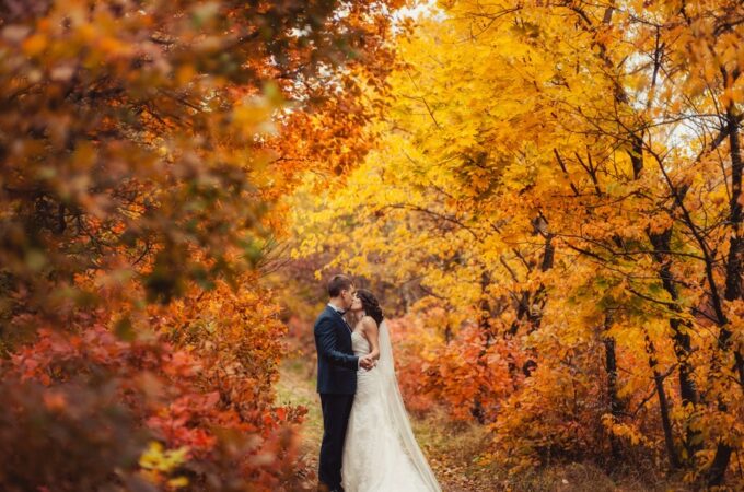 A couple in wedding attire standing close together among orange and yellow autumn trees along a narrow path.