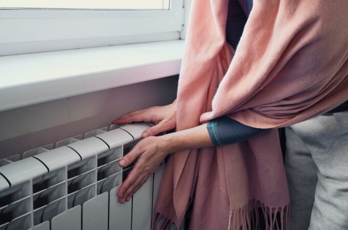 A close-up shot of a person wrapped in a blanket warming their hands over an indoor radiator near a window.