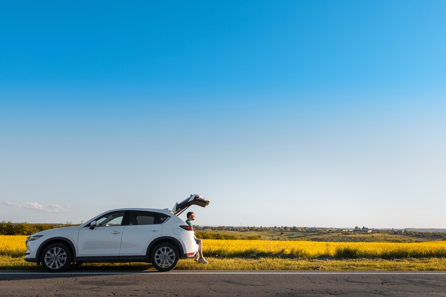 A white car parked on the side of the road on a sunny day. A person sits at the edge of the car’s trunk.