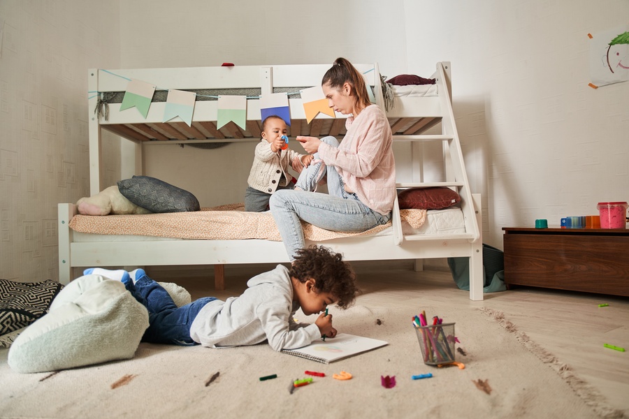 A small child coloring on their bedroom floor as their mom and baby sibling sit on the bottom bunk bed.