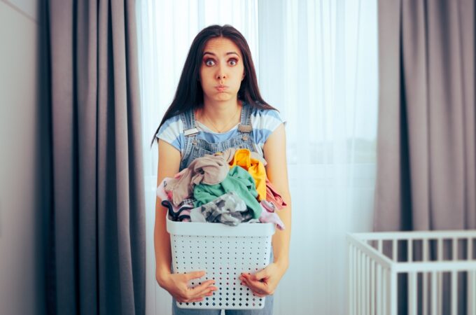 a tired woman holding a full laundry basket in front of curtains, with a white crib visible beside a window.