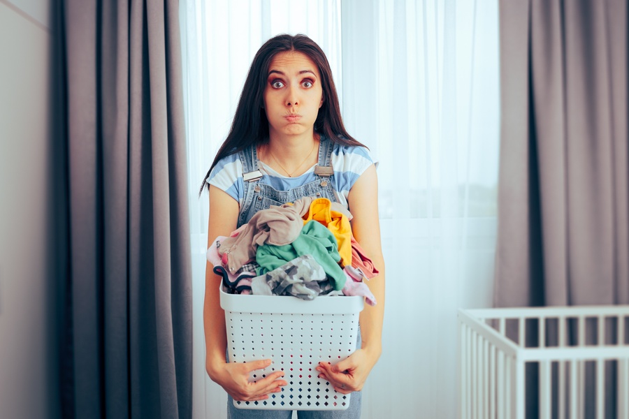 a tired woman holding a full laundry basket in front of curtains, with a white crib visible beside a window.
