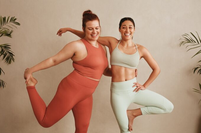 Two women in athletic wear smiling while balancing on one leg in front of a beige wall with plants at the sides.