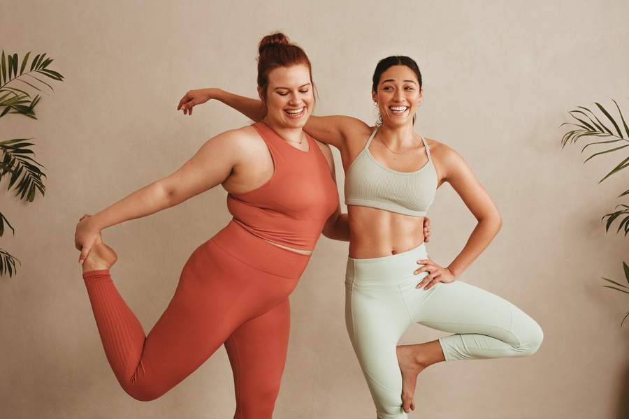 Two women in athletic wear smiling while balancing on one leg in front of a beige wall with plants at the sides.