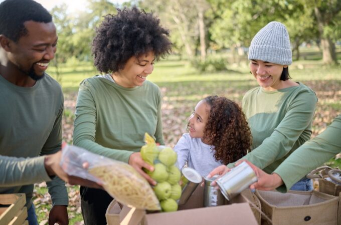 A group of people and a child sort fruits and vegetables into cardboard boxes on a park table under trees.