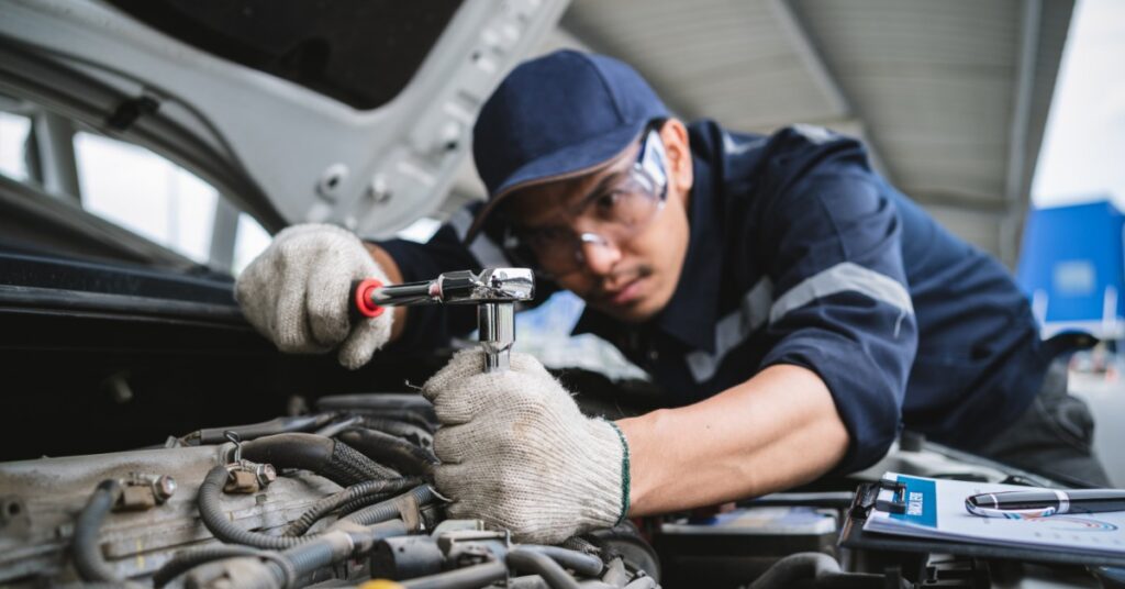 An automotive mechanic with his hands under the vehicle's hood as he unscrews a bolt with a wrench and wears gloves.