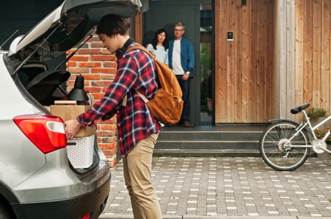 A man wearing a backpack is placing boxes into the trunk of an SUV. His parents are standing on the porch in the background.