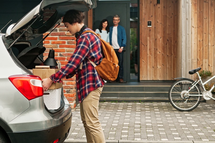  A man wearing a backpack is placing boxes into the trunk of an SUV. His parents are standing on the porch in the background.
