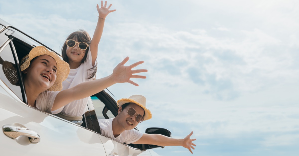 Two adults and a child are leaning out of a white car window, smiling and waving under a bright, cloudy sky.