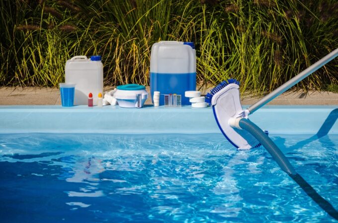 A pool vacuum and hose being held over blue water with chemical containers and testing supplies lined up along the pool edge.