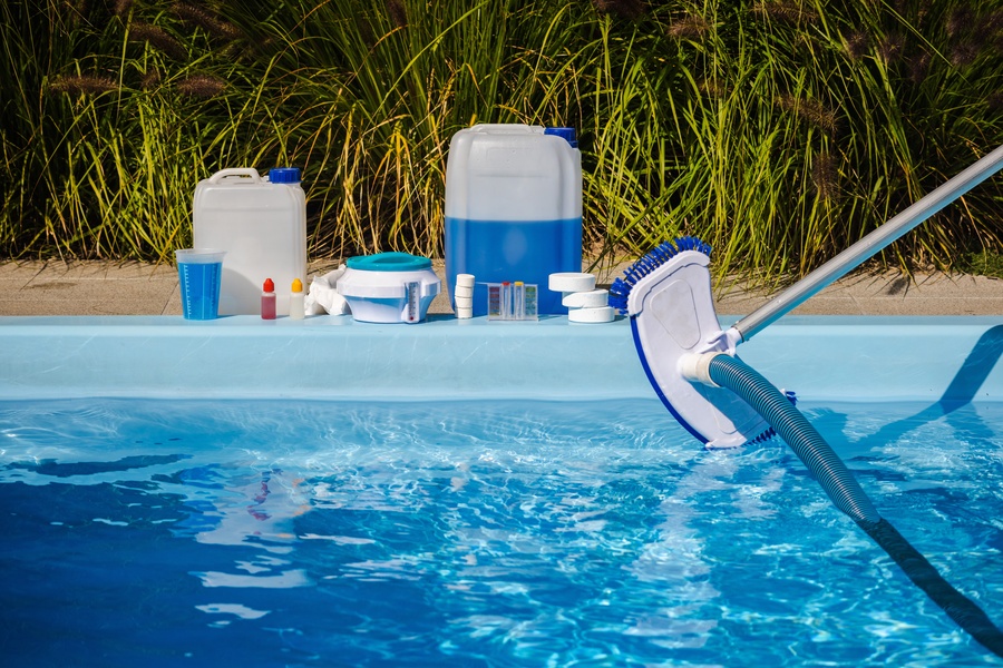 A pool vacuum and hose being held over blue water with chemical containers and testing supplies lined up along the pool edge.