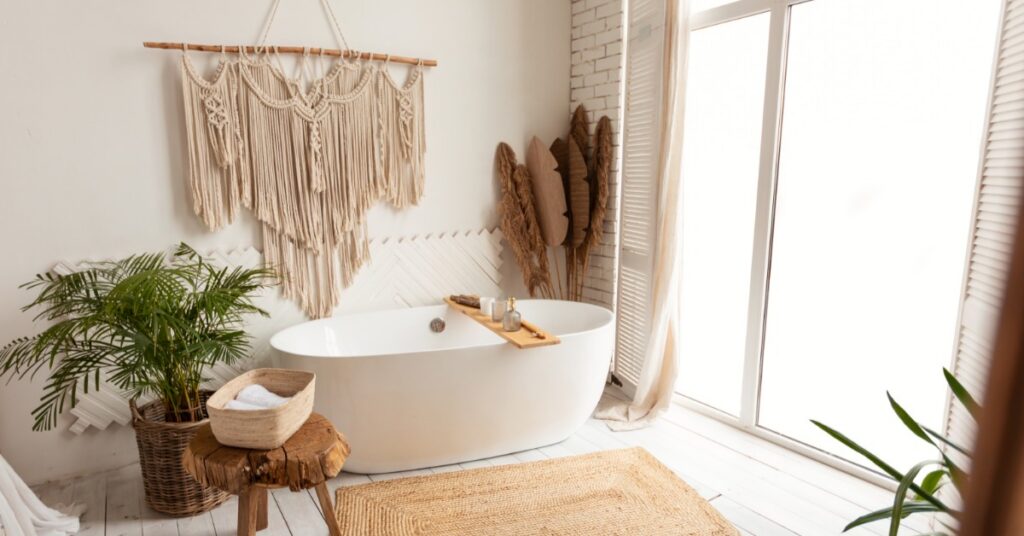 Bright spa-like bathroom with a freestanding tub, large windows, woven rug, wood stool, wall hanging, and plants.
