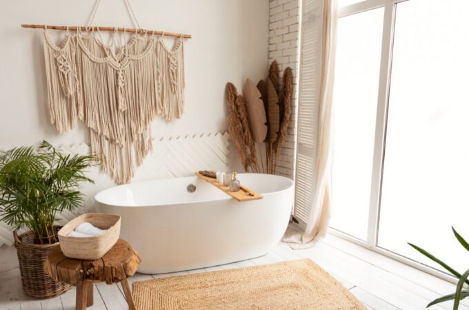 Bright spa-like bathroom with a freestanding tub, large windows, woven rug, wood stool, wall hanging, and plants.