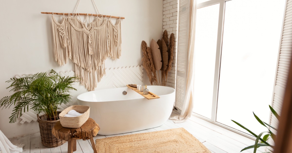 Bright spa-like bathroom with a freestanding tub, large windows, woven rug, wood stool, wall hanging, and plants.