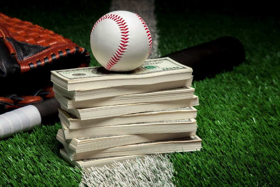 A close-up of a baseball sitting on a stack of dollar bills on a grass field with a bat and glove behind it.