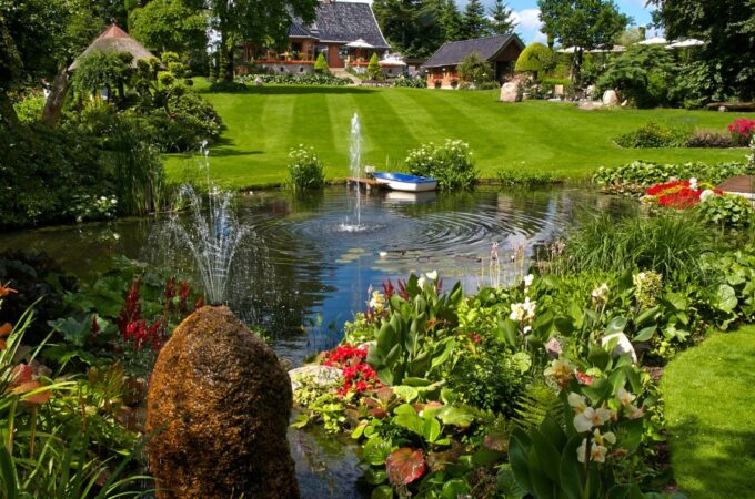 A cute backyard pond features two fountains, beautiful flowers surrounding the edge, and a dock with a small boat.