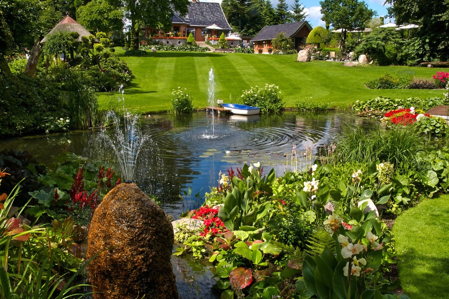 A cute backyard pond features two fountains, beautiful flowers surrounding the edge, and a dock with a small boat.