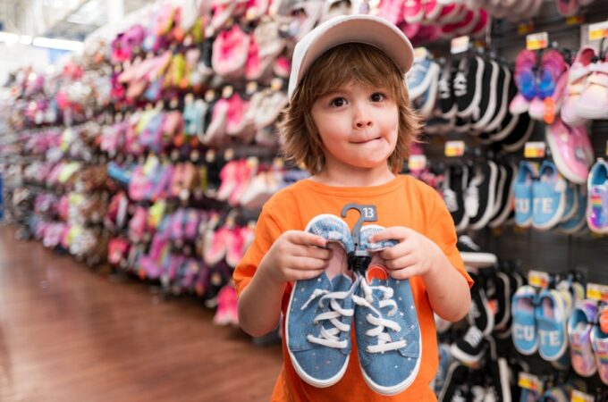 A child in an orange shirt and white hat holding blue sneakers in a store aisle lined with colorful shoes.