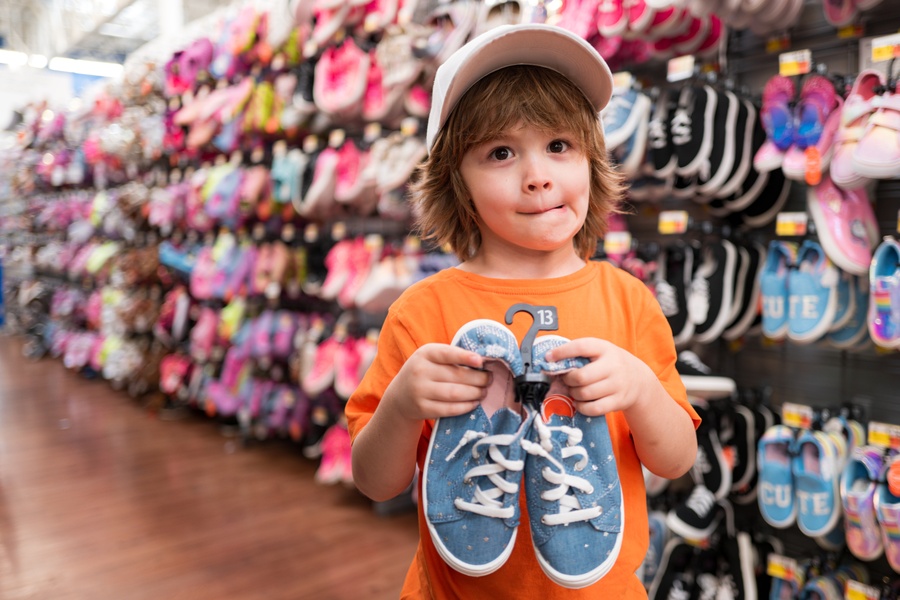 A child in an orange shirt and white hat holding blue sneakers in a store aisle lined with colorful shoes.