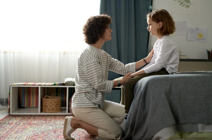 A woman kneeling beside a bed and holding a child's arms while they face each other in a bright bedroom.